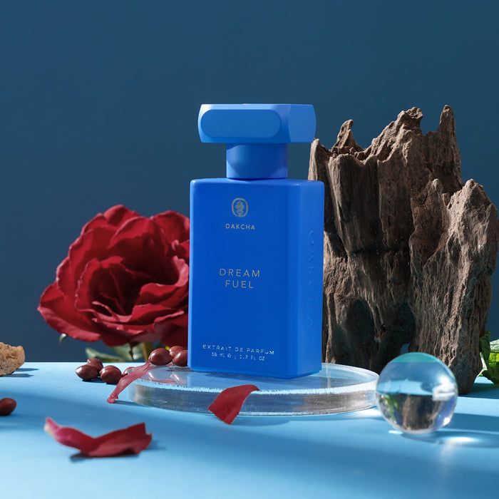 Front view of Dream Fuel blue rectangular perfume bottle showcasing gold lettering, with a red rose and textured wood on a blue background