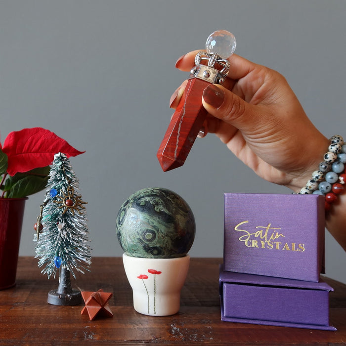 Hand holding a red stone with a crystal ball, surrounded by decorative items on a wooden surface.