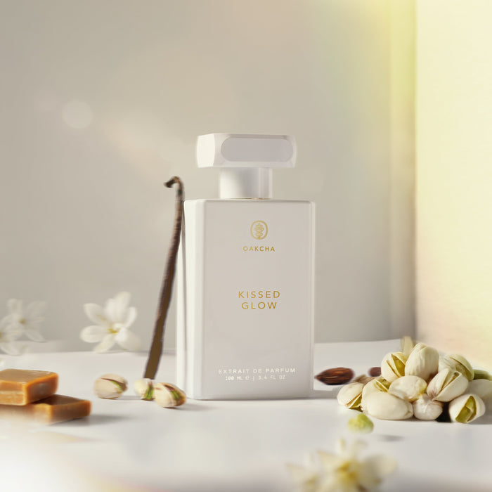 Front view of Kissed Glow 100ml perfume bottle with gold label, surrounded by pistachios and flowers on warm beige background