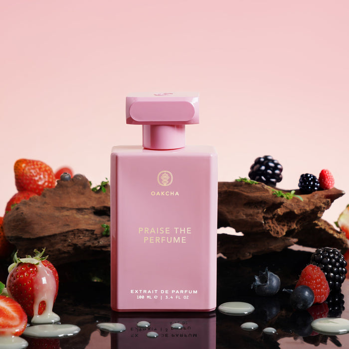 Front view of Praise the Perfume 100mL bottle with gold Oakcha logo, surrounded by fruit and chocolate on reflective surface