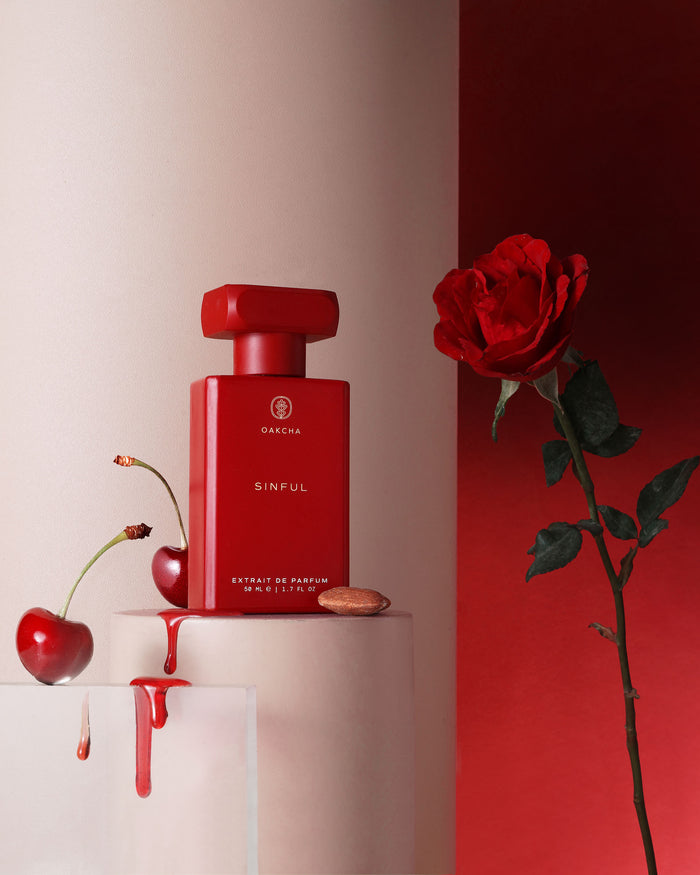 Front view of Sinful Extrait de Parfum showcasing its red bottle with cherries, a rose, and almond on sculptural pedestals