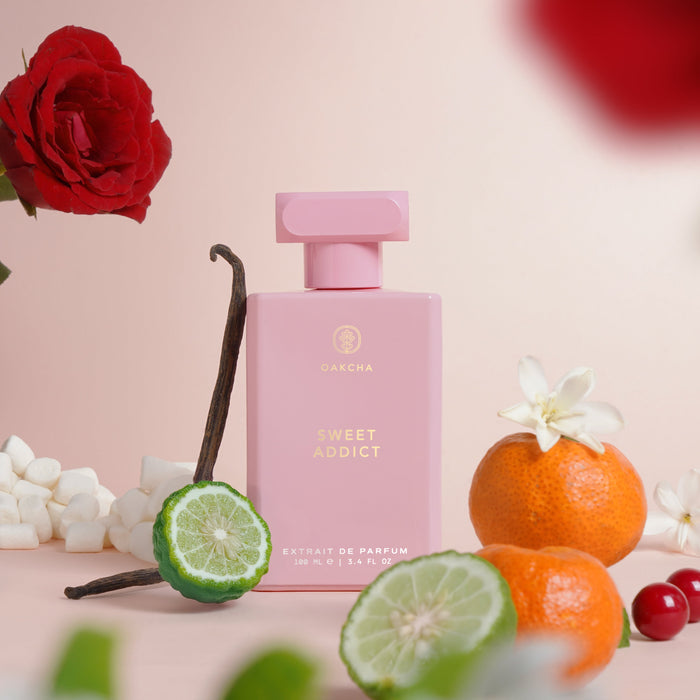 Front view of Sweet Addict 100ml perfume bottle, pink rectangular bottle with cap, citrus and vanilla props on pink background