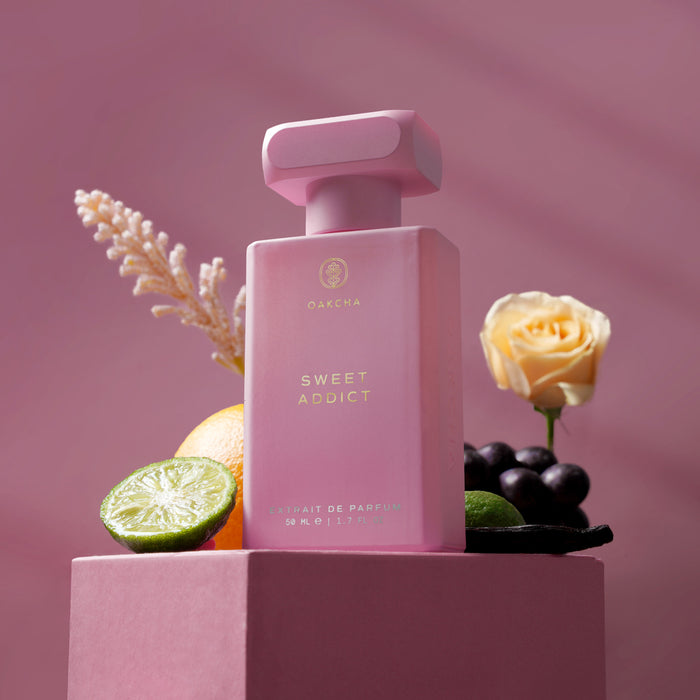 Front view of Sweet Addict perfume showing frosted pink bottle with gold lettering on a pink pedestal with lime and rose