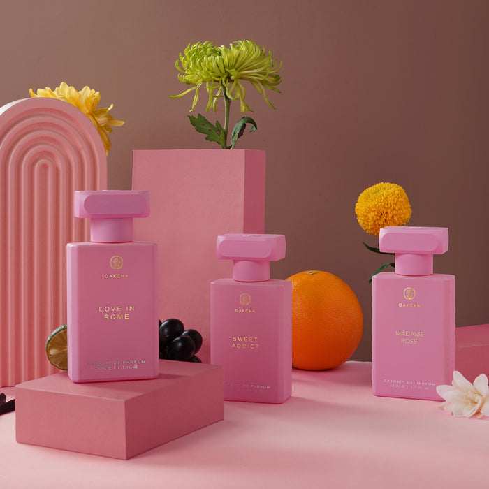 Front view of Sweet Addict perfume bottle in a trio of pink Extrait de Parfum bottles, with flowers and fruit on a pink backdrop