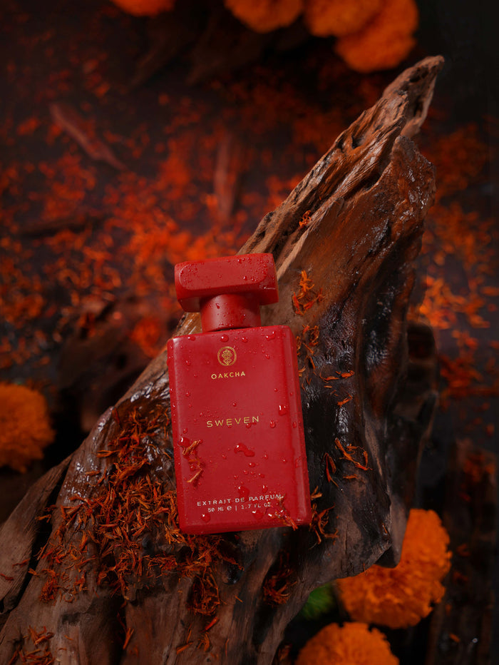 Front view of Sweven 50 milliliter extrait de parfum bottle showcasing its red finish and water droplets on dark driftwood with orange flowers